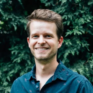 Smiling man with light brown hair wearing a dark blue patterned shirt against a background of green foliage.